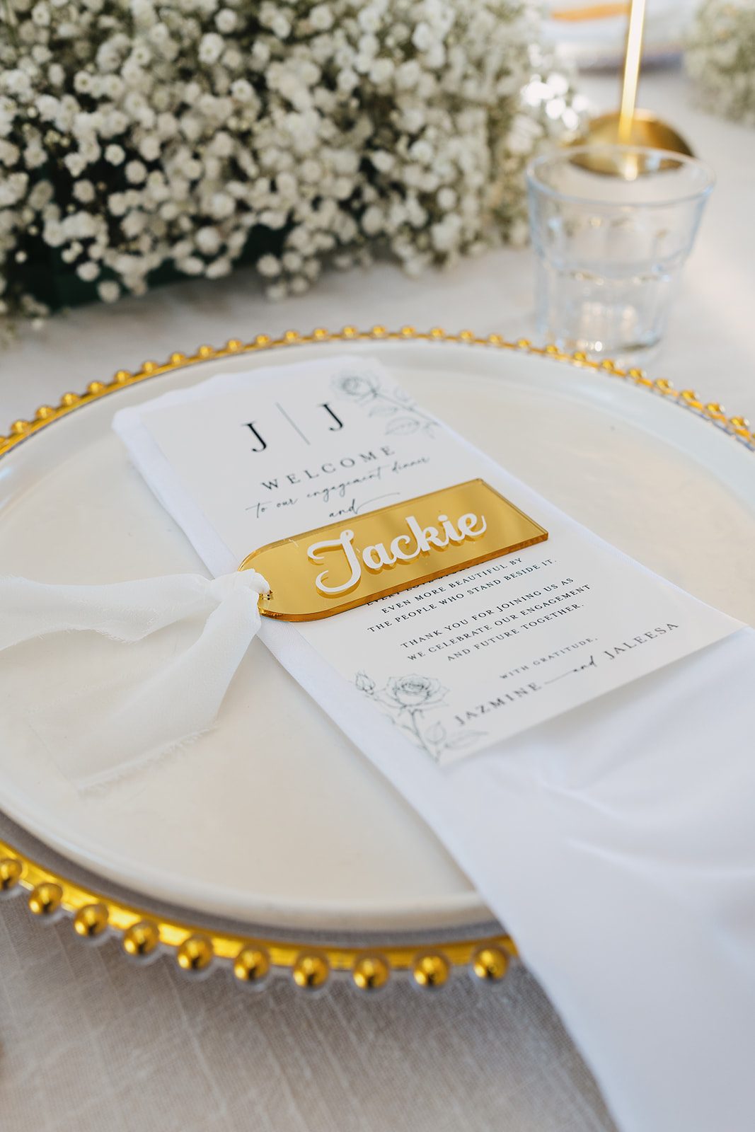 A white welcome menu with a gold acrylic name card resting on a gold-beaded charger, with baby's breath and glassware softly blurred behind