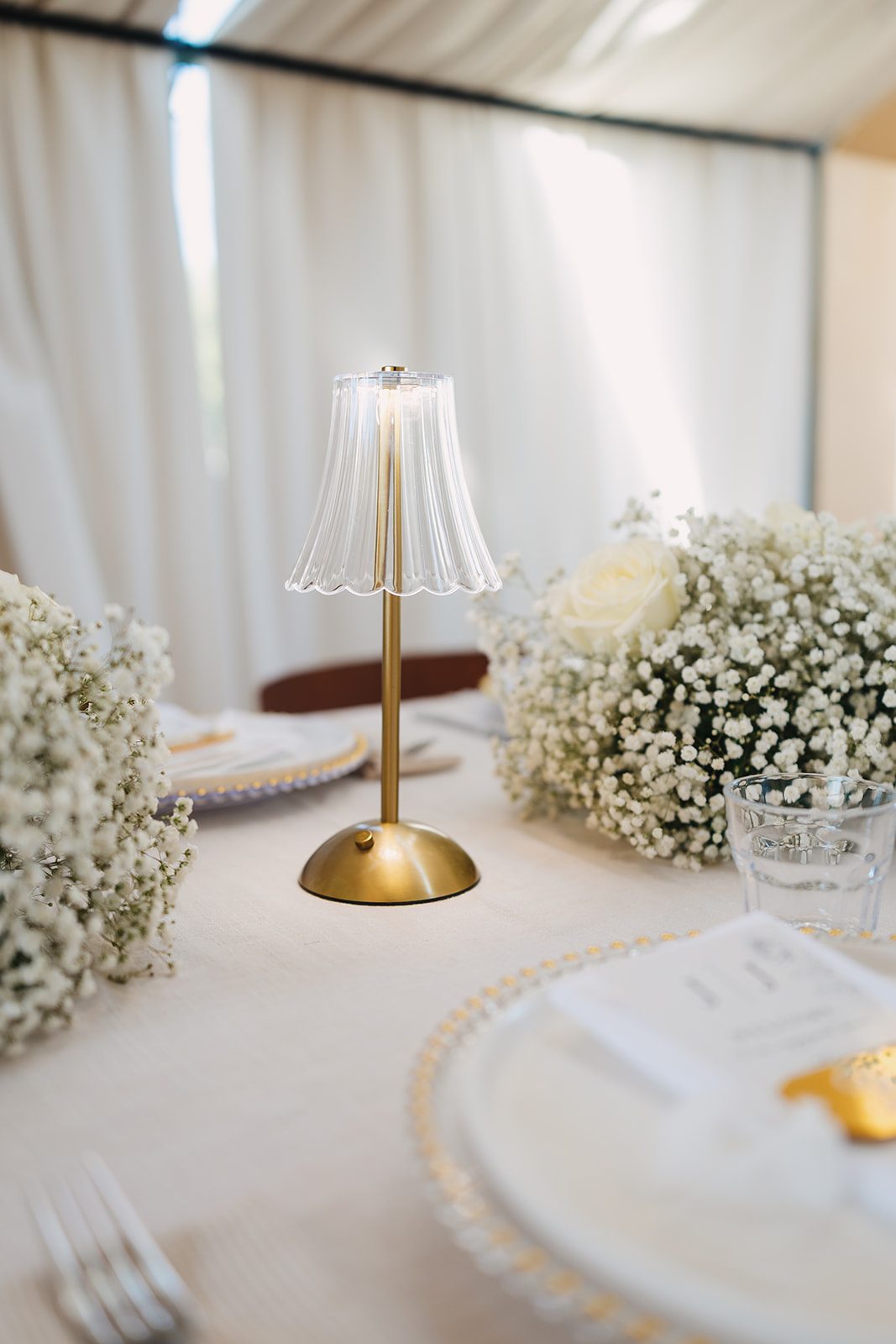 A gold cordless lamp with a fluted glass shade between low clusters of baby's breath and a white rose, on a long white-draped table set with gold-beaded chargers, under a sheer white tent