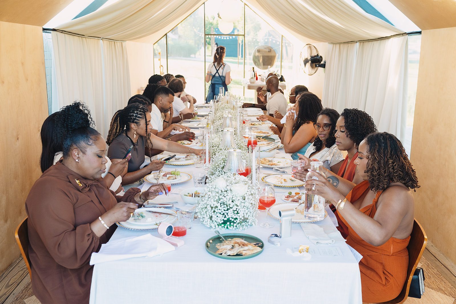 Guests seated along a long white-draped table under a sheer white tent, with baby's breath florals, gold-rimmed place settings and glassware down the center