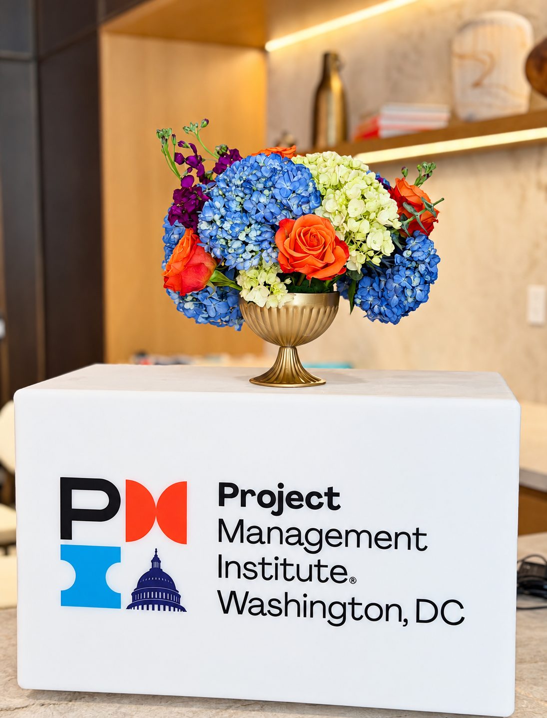 Blue hydrangeas and orange roses in a gold pedestal bowl on a white plinth branded Project Management Institute Washington, DC Chapter