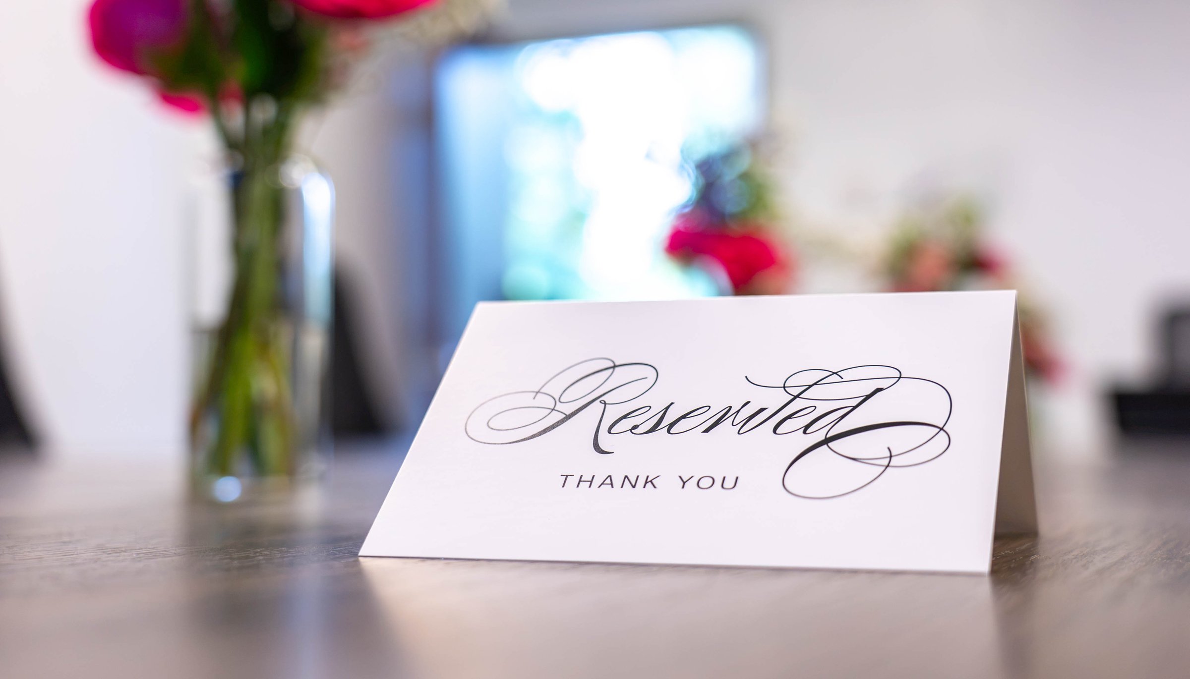 A white folded card reading Reserved in black calligraphy with Thank You beneath it, on a wood table in front of pink florals at the Able Health Services grand opening