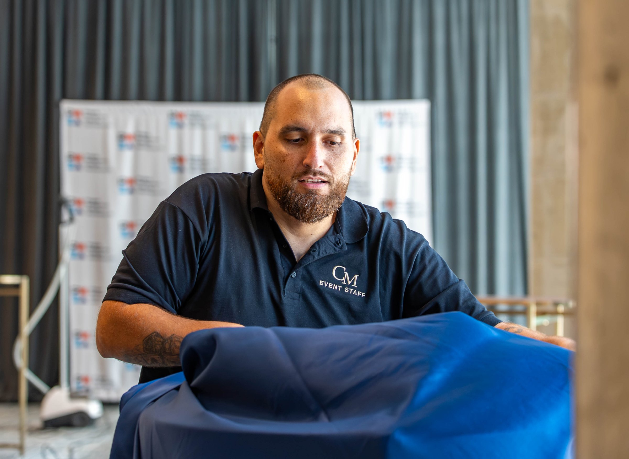 A Creative Motion Events team member unfolding a navy linen during setup, with the sponsor step-and-repeat behind him, at the PMI Washington DC Chapter Awards Gala