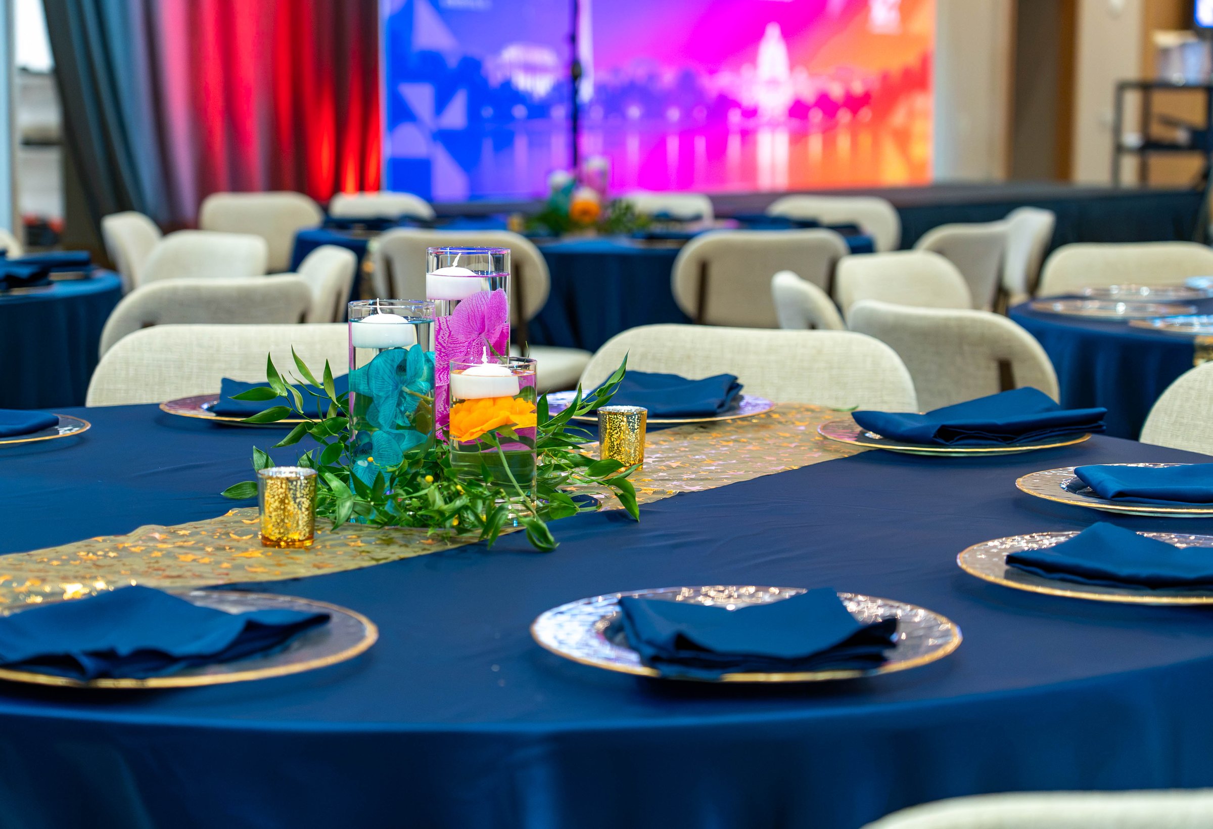 A navy-linen gala table with gold-rimmed chargers, navy napkins, a gold sequin runner and a turquoise and magenta orchid floating-candle centerpiece, with the stage screen glowing magenta and blue behind, at the PMI Washington DC Chapter Awards Gala