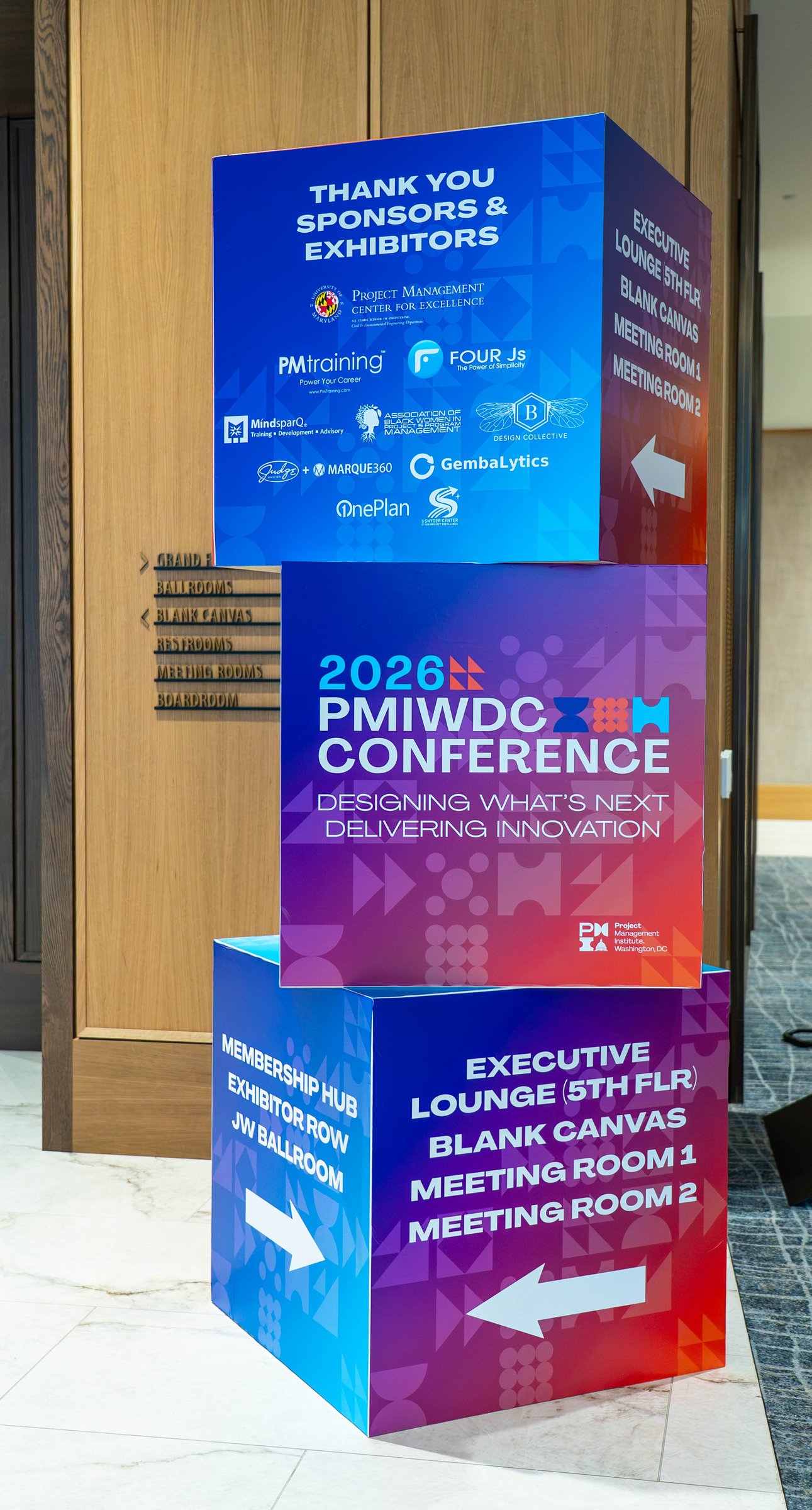 A stacked signage tower of printed cubes reading Thank You Sponsors & Exhibitors, 2026 PMIWDC Conference, and directions to the Executive Lounge and meeting rooms, at the PMI Washington DC Chapter Awards Gala