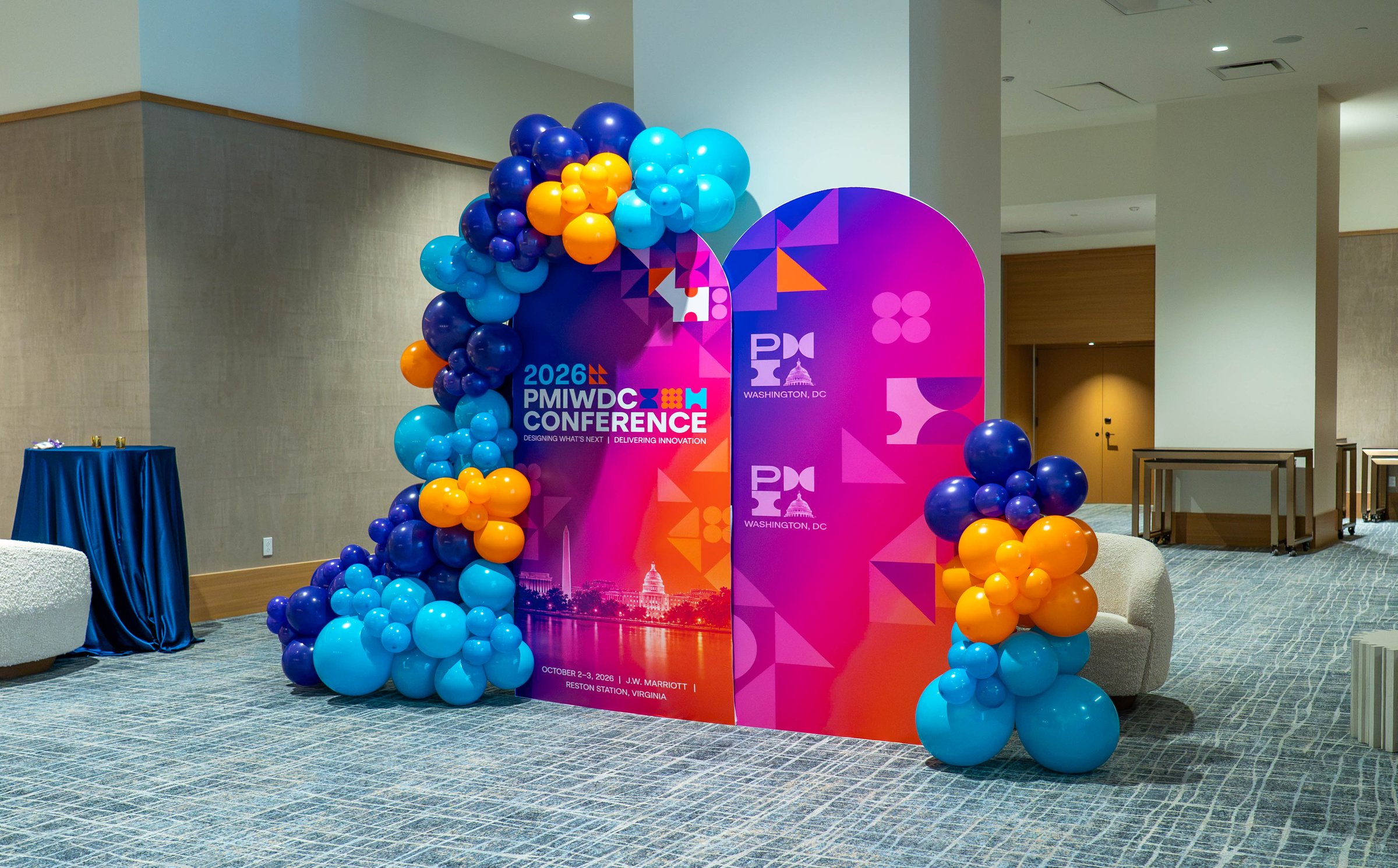 A navy, turquoise and orange balloon arch wrapped around an arched 2026 PMIWDC Conference welcome panel in magenta and blue, in the pre-function area at the PMI Washington DC Chapter Awards Gala