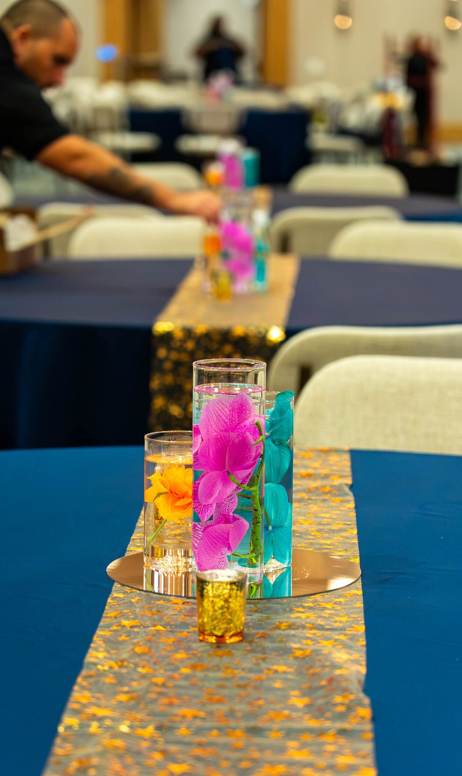 Row of orchid centerpieces on gold runners while the team sets tables for the PMI Washington DC Chapter Awards Gala, JW Marriott Reston, Virginia