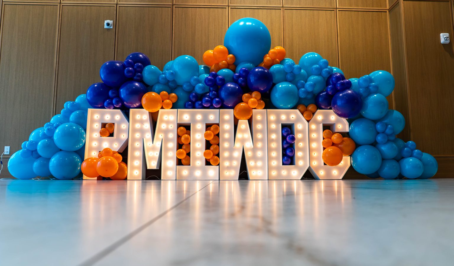 Illuminated PMIWDC marquee letters framed by a blue and orange balloon garland